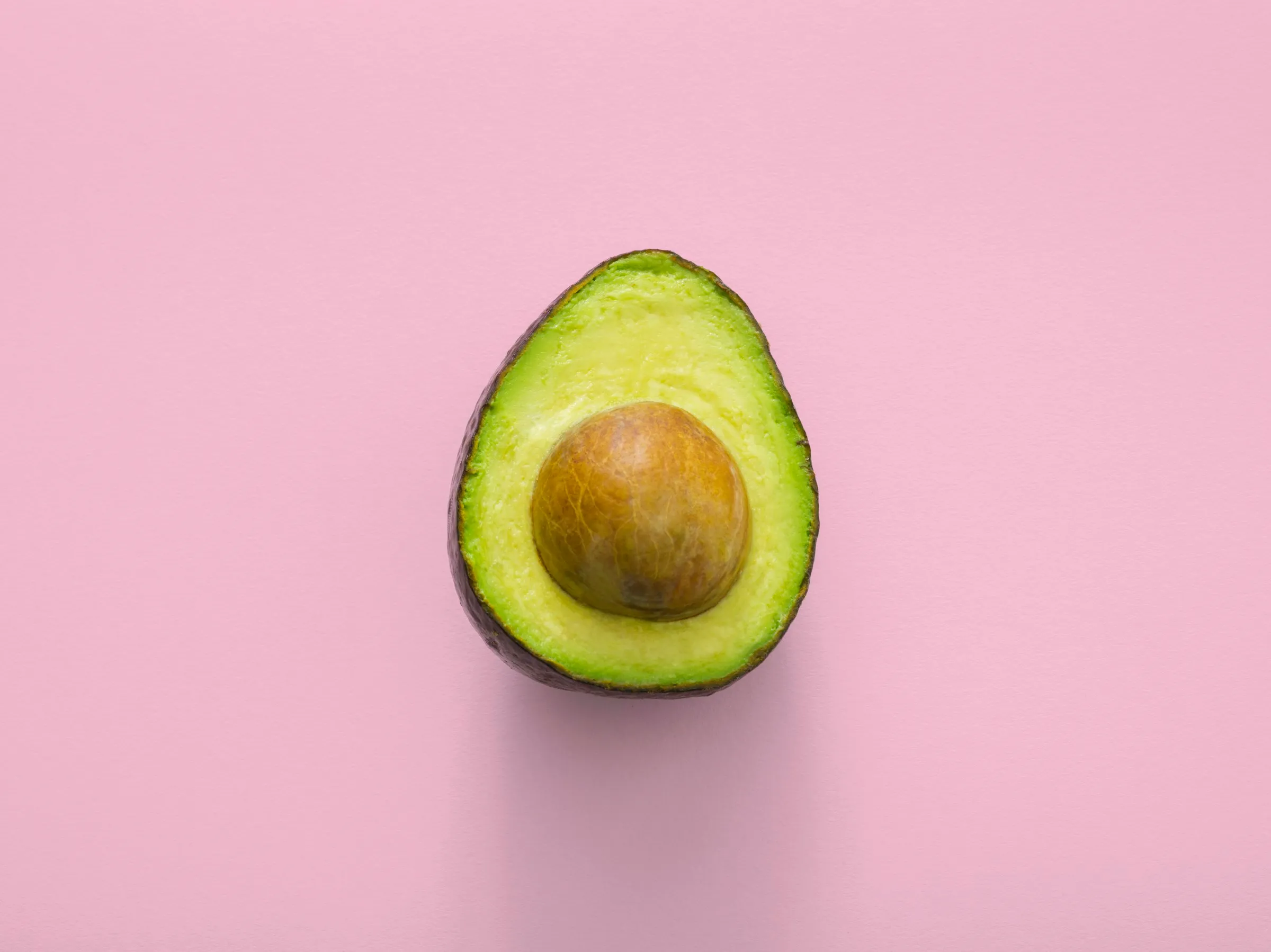 A perfectly ripe avocado sliced in half, showing its creamy green flesh and round brown pit, photographed from above on a soft pink background.
