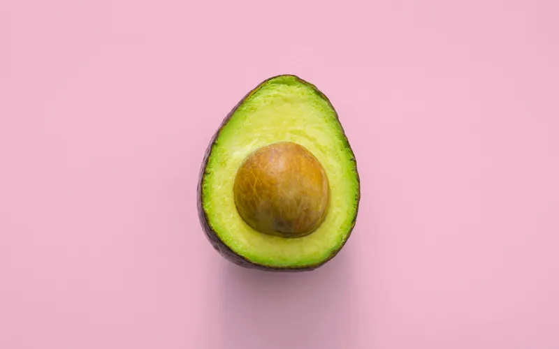A perfectly ripe avocado sliced in half, showing its creamy green flesh and round brown pit, photographed from above on a soft pink background.