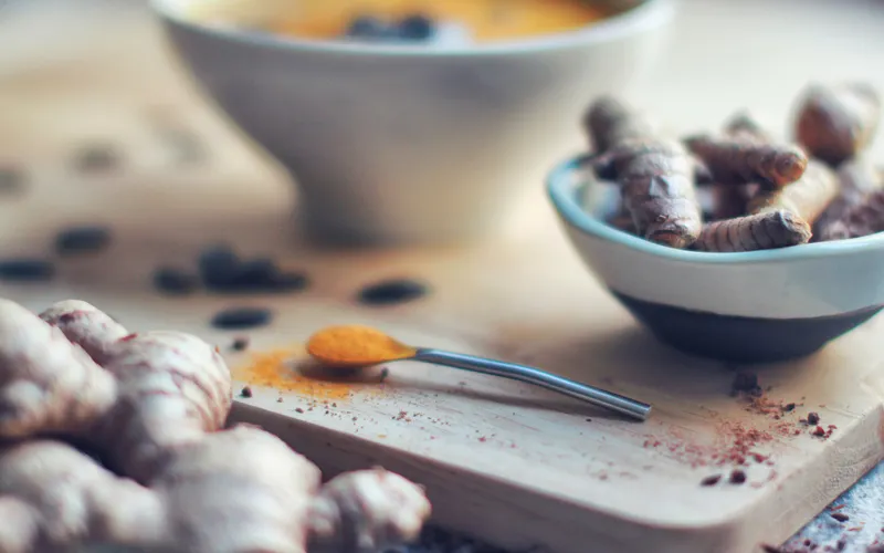 A warm golden turmeric latte beside a small bowl of fresh turmeric roots and a spoon heaped with vivid orange turmeric powder, with whole peppercorns and fresh ginger scattered on a wooden board