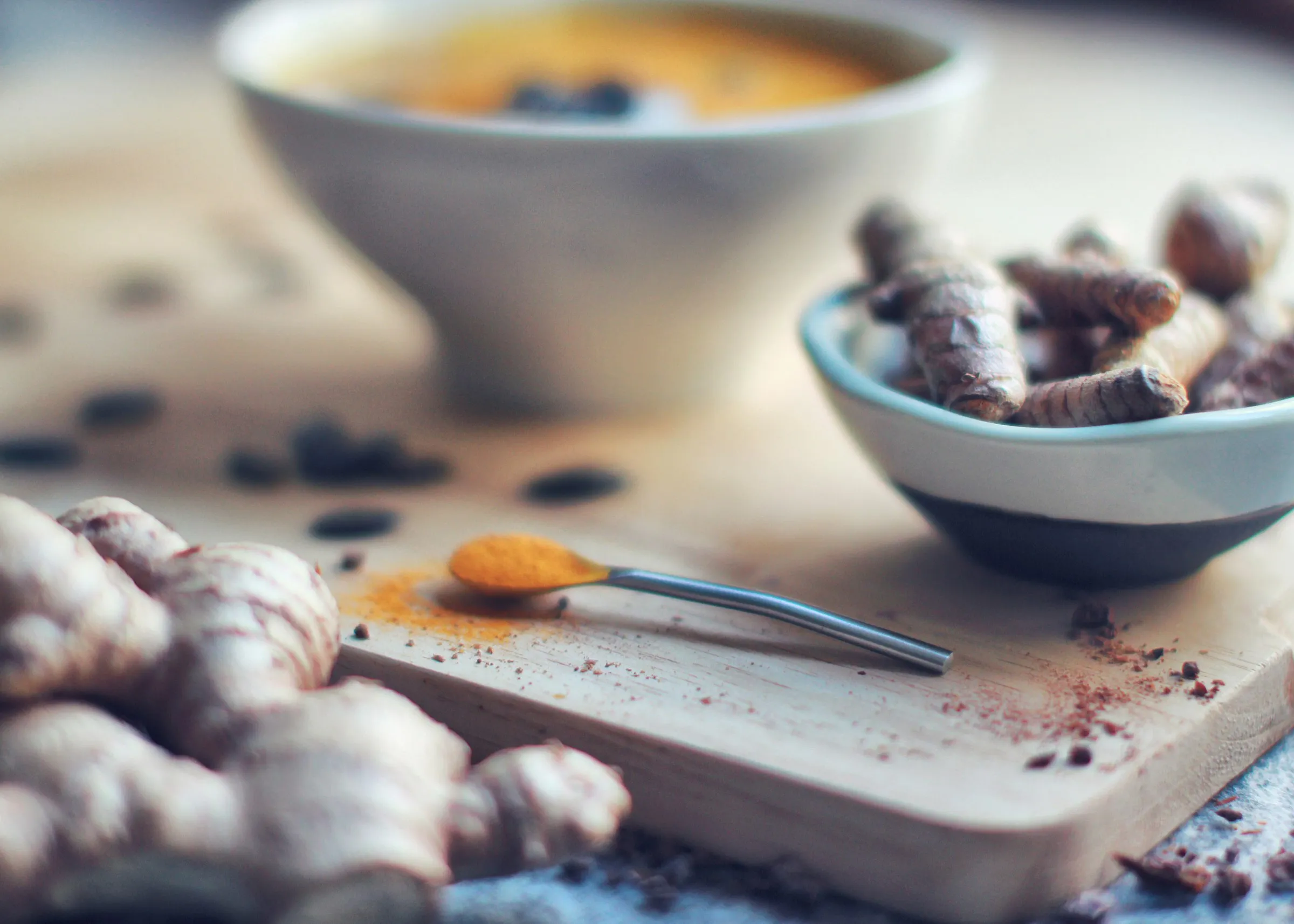 A warm golden turmeric latte beside a small bowl of fresh turmeric roots and a spoon heaped with vivid orange turmeric powder, with whole peppercorns and fresh ginger scattered on a wooden board